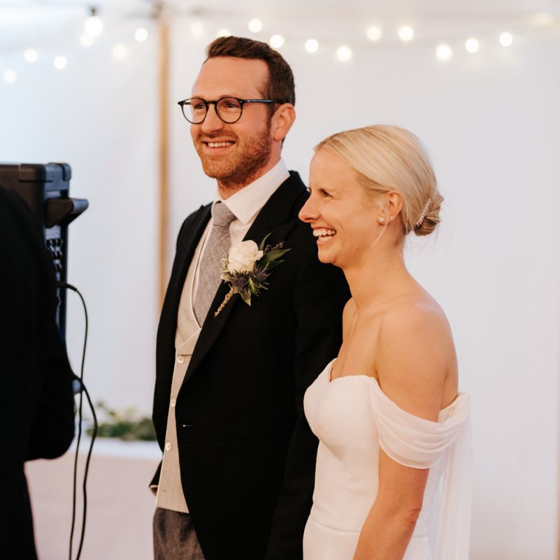Bride and groom laugh and smile, holding hands, while fathe of the bride delivers wedding speech