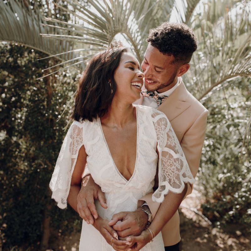 Groom in pale pink blazer embracing his boho bride in a Grace Loves Lace wedding dress