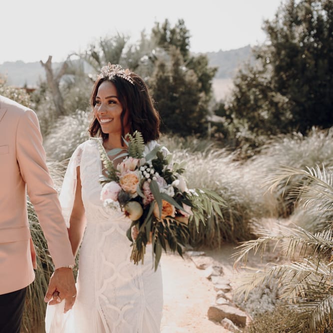 Groom in pink blazer and floral bow tie holding his brides hand in Spain