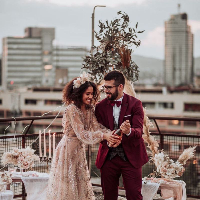 Bride in sparkle wedding dress and groom in burgundy suit popping champagne at rooftop wedding