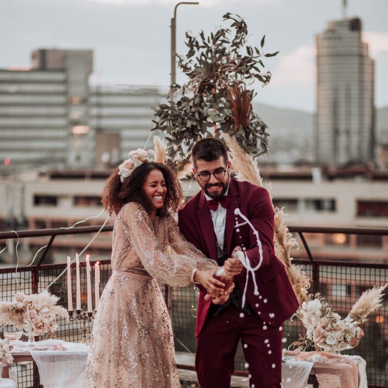 Bride in sparkle wedding dress and groom in burgundy suit popping champagne