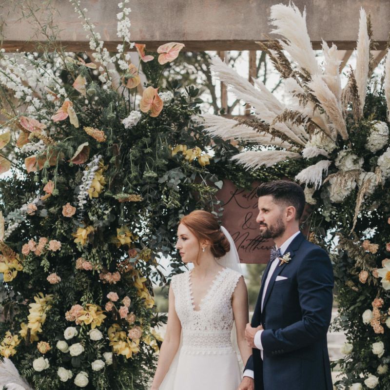 Bride in Pronovias Wedding Dress and Groom in Navy Suit Holding Hands in Front of a Peach Floral Arch