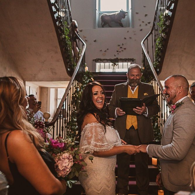 Bride in Off the Shoulder Grace Loves Lace Wedding Dress and Groom in Grey Blazer and Bow Tie Holding Hands at the Wedding Ceremony