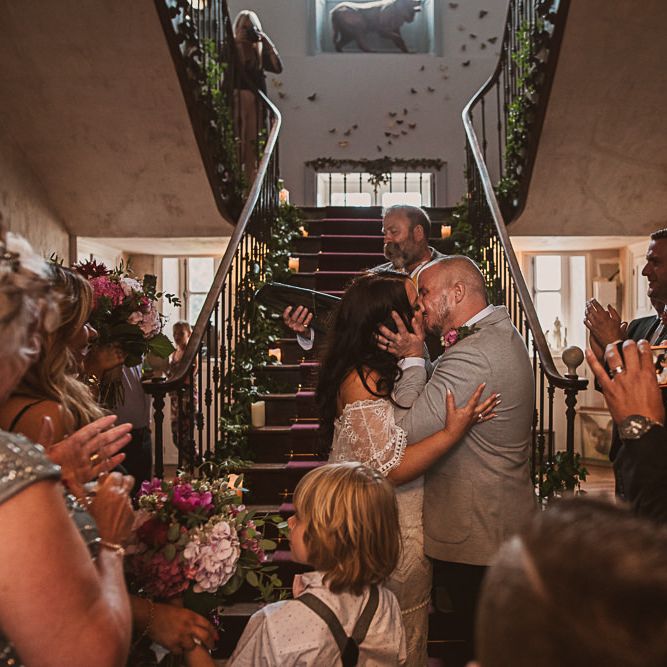 Bride in Off the Shoulder Grace Loves Lace Wedding Dress and Groom in Grey Blazer and Bow Tie Kissing at the Wedding Ceremony