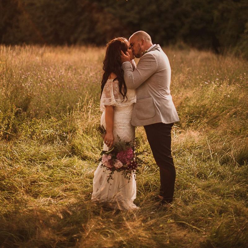 Bride in Emanuela Grace Loves Lace Wedding Dress and Groom in Grey Blazer Kissing in a Field