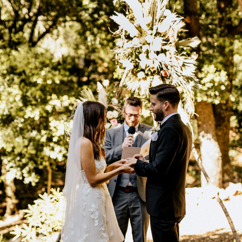 Bride in Tiered Lace Emma Beaumont Wedding Dress and Groom in Givenchy Tuxedo Exchanging Vows
