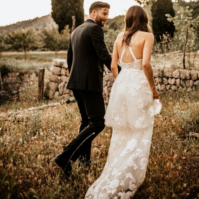 Bride in Tiered Lace Emma Beaumont Wedding Dress and Groom in Givenchy Tuxedo Walking Through Fields