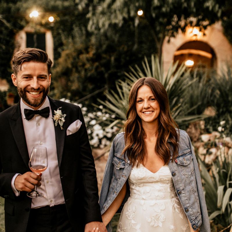 Bride in Tiered Lace Emma Beaumont Wedding Dress and Denim Jacket and Groom in Givenchy Tuxedo Laughing Whilst Holding Hands