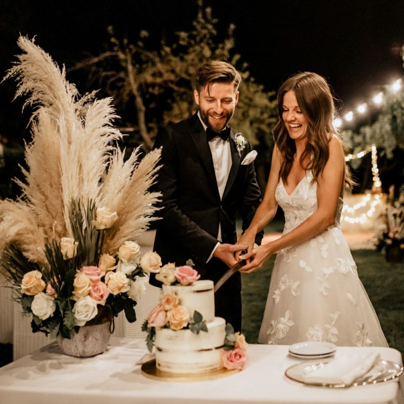 Bride in Tiered Lace Emma Beaumont Wedding Dress and Denim Jacket and Groom in Givenchy Tuxedo Cutting the Cake