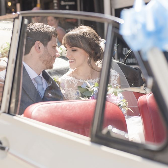 Bride and Groom in White Morris Minor with Blue Ribbon and Flowers | Bride in Lacy Fishtail Stella York Gown with Plunging V Back and Diamante Charm on Champagne Ribbon Belt | Summer Bridal Bouquet with Purple and Blue Flowers and Eucalyptus | Groom in Blue Jacket and Waistcoat with Camel Chinos | Morris Minor Wedding Car, Hanging Flowers in Bottles and Town Crier for Village Hall Wedding in Rye | Eleanor Jane Photography