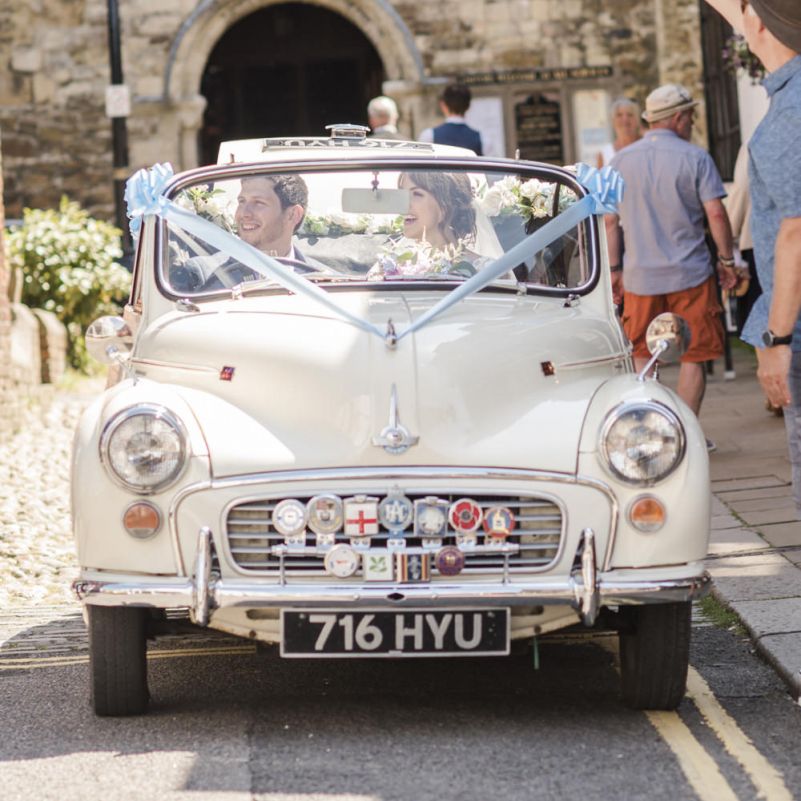 Bride and Groom in White Morris Minor with Blue Ribbon and Flowers | Bride in Lacy Fishtail Stella York Gown with Plunging V Back and Diamante Charm on Champagne Ribbon Belt | Summer Bridal Bouquet with Purple and Blue Flowers and Eucalyptus | Groom in Blue Jacket and Waistcoat with Camel Chinos | Morris Minor Wedding Car, Hanging Flowers in Bottles and Town Crier for Village Hall Wedding in Rye | Eleanor Jane Photography