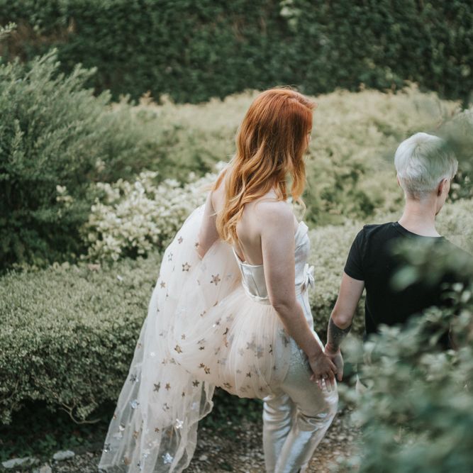 Bride in jumpsuit with gold star covered train walking through the gardens at Castle Combe Manor House in Wiltshire