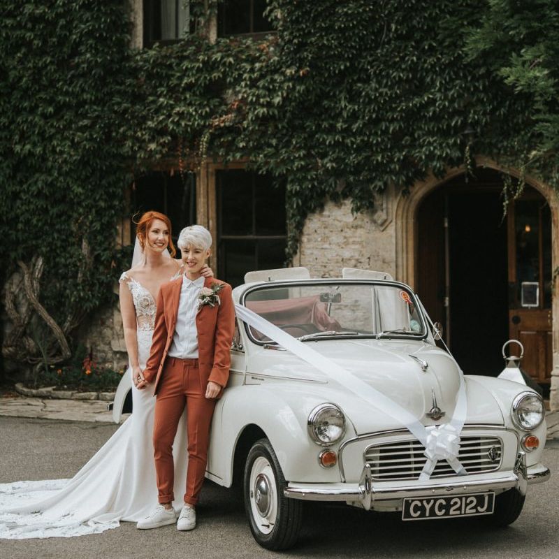 Two brides standing next to a morris Minor wedding cake