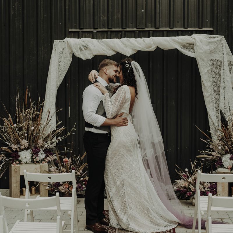 Boho Bride in Lace Wedding Dress and Groom Kissing at the Altar with Drapes, Woven Rug, Bold Sign and Dried Flower Arrangements