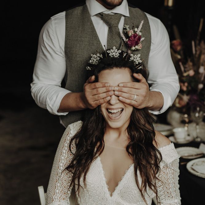 Groom in Wool Waistcoat covering Brides Eyes  in Lace Wedding Dress and Crown Headpiece