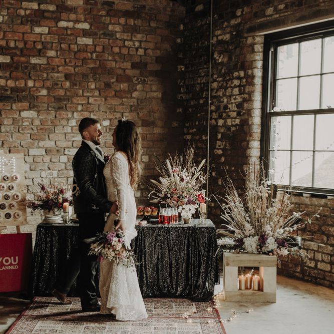 Boho Bride in Lace Wedding Dress and Groom in Leather Jacket Standing at Dessert Table with Doughnut Wall, Wedding Cakes and Dried Flower Arrangements