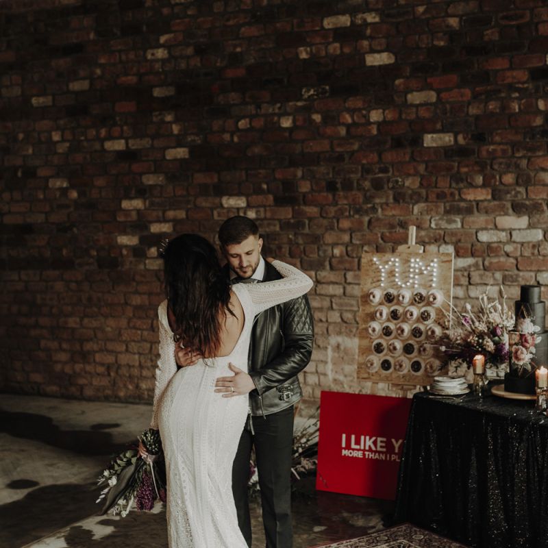 Boho Bride in Lace Wedding Dress with Low Back and Groom in Leather Jacket Embracing Next to the Dessert Table