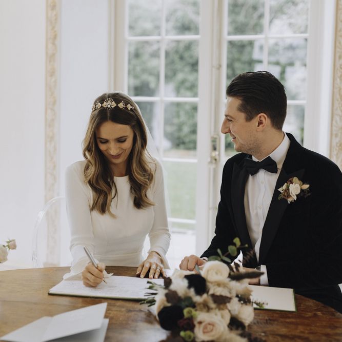 Bride in Emma Beaumont Wedding Dress and Tilly Thomas Lux Headdress and Groom in Black Tie Suit Signing the Register