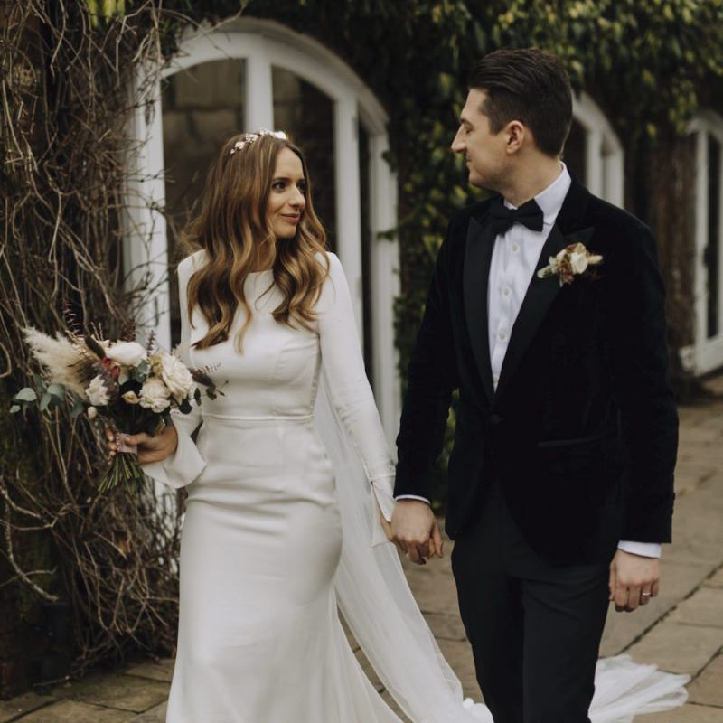 Bride in Emma Beaumont Wedding Dress and Groom in Tuxedo Walking Hand in Hand