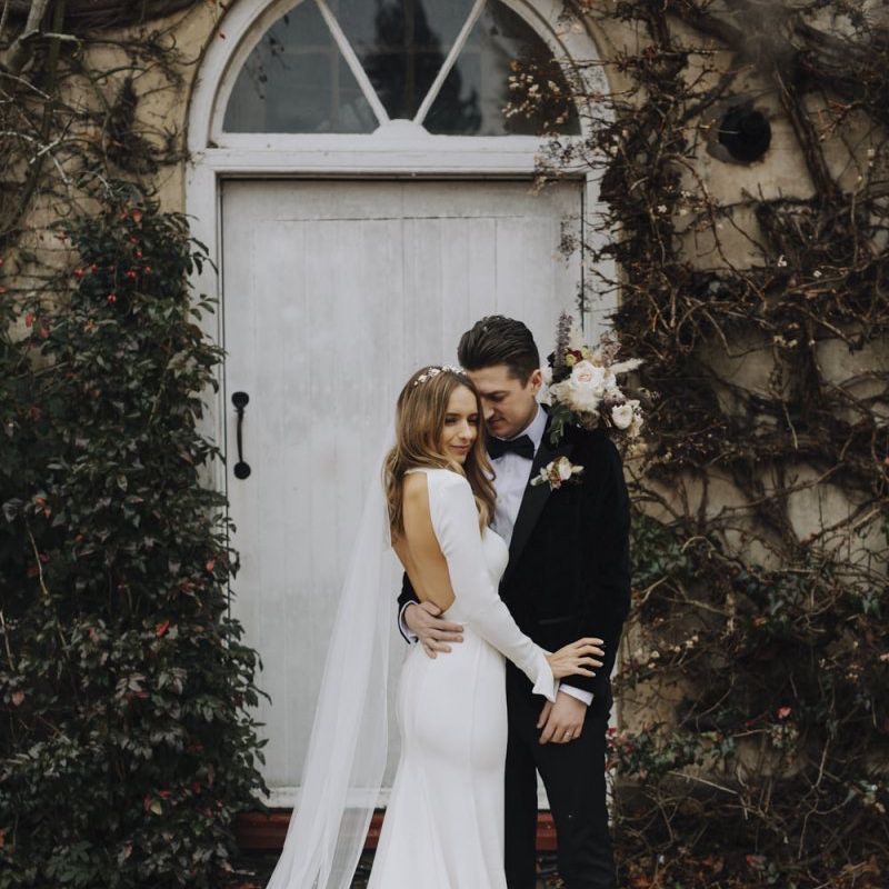 Bride in Backless Emma Beaumont Wedding Dress and Groom in Tuxedo Embracing