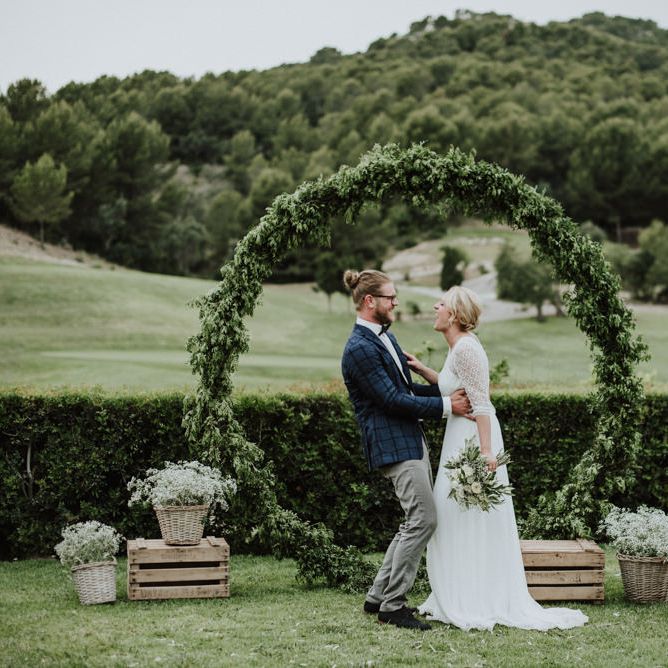 Bride in Rembo Styling Wedding Dress and Groom  in Chino's and Checked Blazer Standing in Front of A Greenery Moon Gate