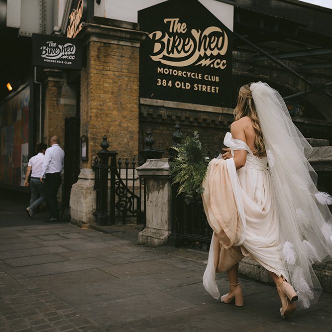 Bride Walking to Wedding Ceremony at The Bike Shed Motorcycle Club in Shoreditch | Bespoke Wedding Gown with Blush Underskirt and One Off Shoulder Strap | Fingertip Length Veil Customised with Feathers | Half Up Half Down Bridal Hair | Nude Topshop Shoes with Pom Pom | Homemade Bouquet of White Flowers, Ferns and Foliage | Bike Shed Motorcycle Club Wedding for ELLE Digital Editor | Nigel John Photography