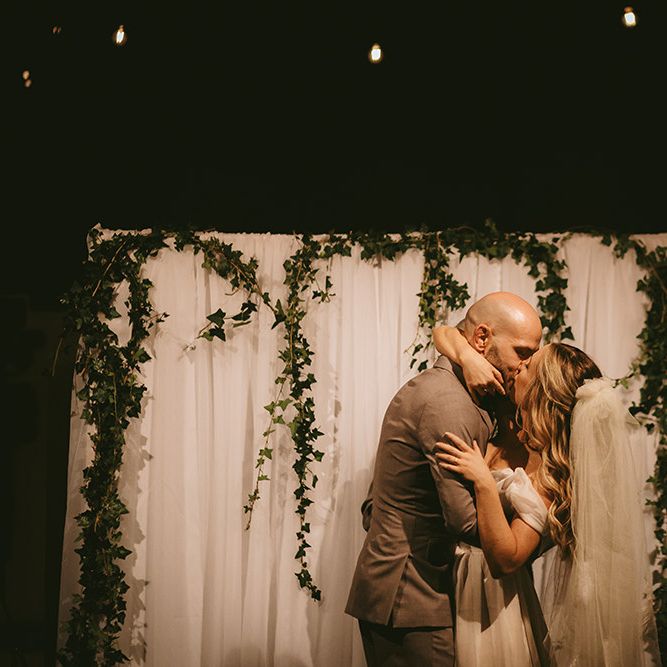 First Kiss | Altar with White Drapery and Foliage Garland | Bride in  Bespoke Wedding Gown with Blush Underskirt and One Off Shoulder Strap | Fingertip Length Veil Customised with Feathers | Groom in Grey Puppy Tooth Check Two-Piece Paul Smith Suit with Amber Tie Pin, Pocket Square and Braces | Bike Shed Motorcycle Club Wedding for ELLE Digital Editor | Nigel John Photography