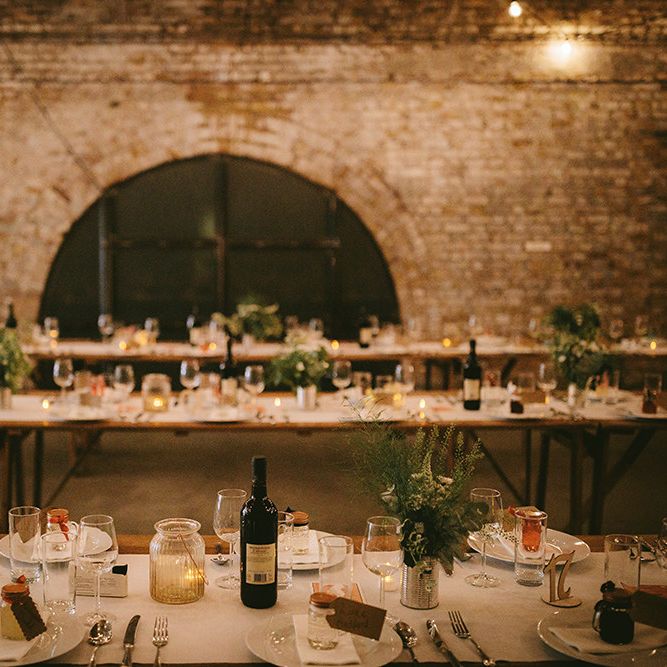 Wedding Reception Décor at The Bike Shed Motorcycle Club in Shoreditch | Wooden Trestle Tables with White Runner | White Flowers, Ferns and Foliage in Tin Can | Tea Lights in Large Textured Jars | Bike Shed Motorcycle Club Wedding for ELLE Digital Editor | Nigel John Photography