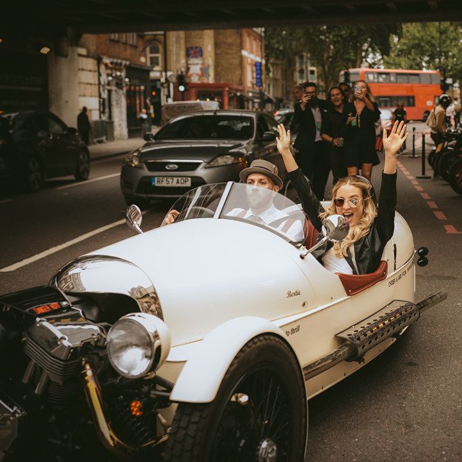 Bride and Groom Taking a Spin in Motorbike and Sidecar | Groom in Grey Puppy Tooth Check Two-Piece Paul Smith Suit with Amber Tie Pin, Pocket Square, Braces and Hat with Feather | Bride in Bespoke Wedding Gown with Blush Underskirt and One Off Shoulder Strap | Thug Wife Leather Bridal Jacket | Bride Wearing Aviators | Bike Shed Motorcycle Club Wedding for ELLE Digital Editor | Nigel John Photography