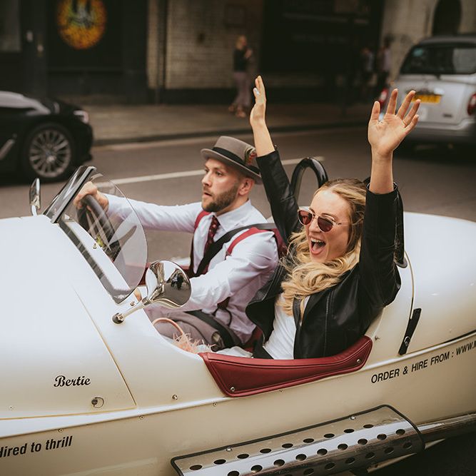 Bride and Groom Taking a Spin in Motorbike and Sidecar | Groom in Grey Puppy Tooth Check Two-Piece Paul Smith Suit with Amber Tie Pin, Pocket Square, Braces and Hat with Feather | Bride in Bespoke Wedding Gown with Blush Underskirt and One Off Shoulder Strap | Thug Wife Leather Bridal Jacket | Bride Wearing Aviators | Bike Shed Motorcycle Club Wedding for ELLE Digital Editor | Nigel John Photography