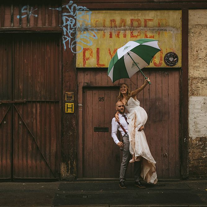 Bride in Bespoke Wedding Gown with Blush Underskirt and One Off Shoulder Strap | Nude Topshop Shoes with Pom Pom | Groom in Grey Puppy Tooth Check Two-Piece Paul Smith Suit with Amber Tie Pin, Pocket Square and Braces | Green and White Umbrella | Bike Shed Motorcycle Club Wedding for ELLE Digital Editor | Nigel John Photography