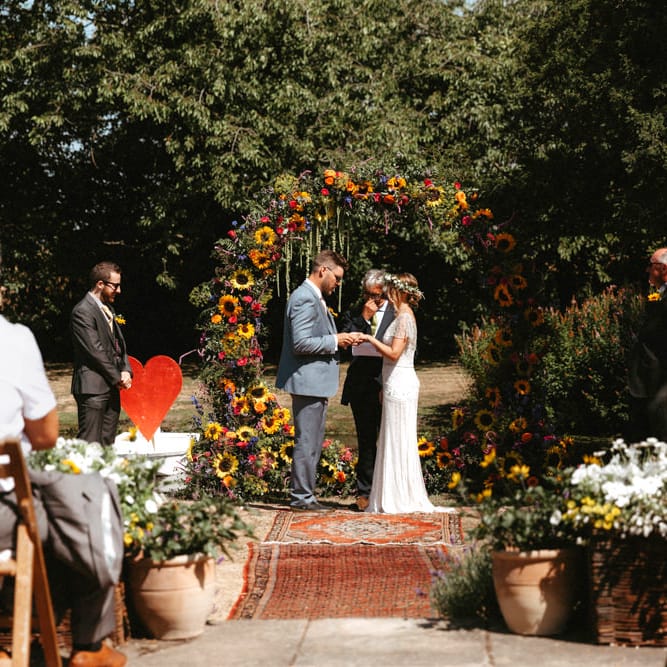 Bride in Jenny Packham Portia Wedding Dress and Groom in Grey Ted Baker Suit Exchanging Vows at Outdoor Wedding Ceremony with Floral Moon Gate,