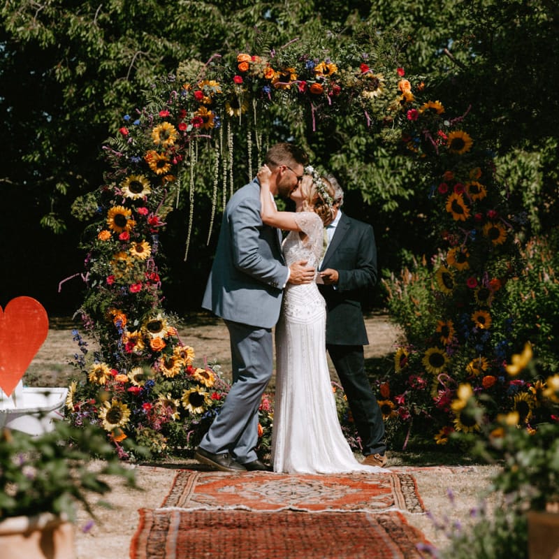 Bride in Jenny Packham Portia Wedding Dress and Groom in Grey Ted Baker Suit Kissing at the Floral Moon Gate Altar
