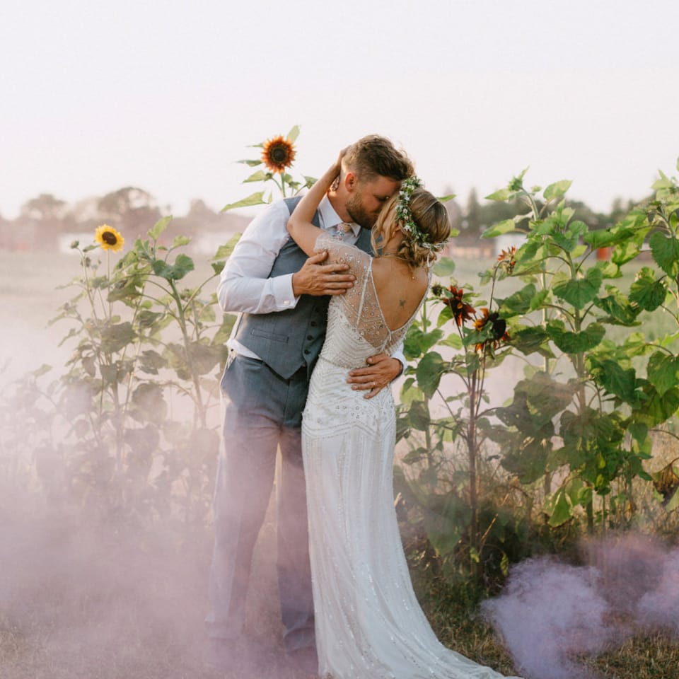Bride in Jenny Packham Portia Wedding Dress and Flower Crown and Groom in Ted Baker Suit Kissing in a Sunflower Field with Smoke Bombs Going Off