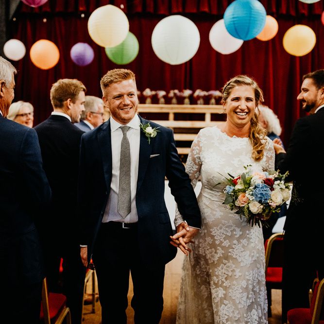 Groom wearing navy two piece with the bride wearing laced long sleeve dress and brightly coloured floral bouquet