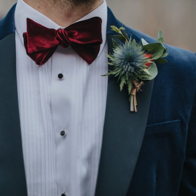 Navy velvet grooms suit details with red bow tie and festive buttonhole foliage for winter wedding