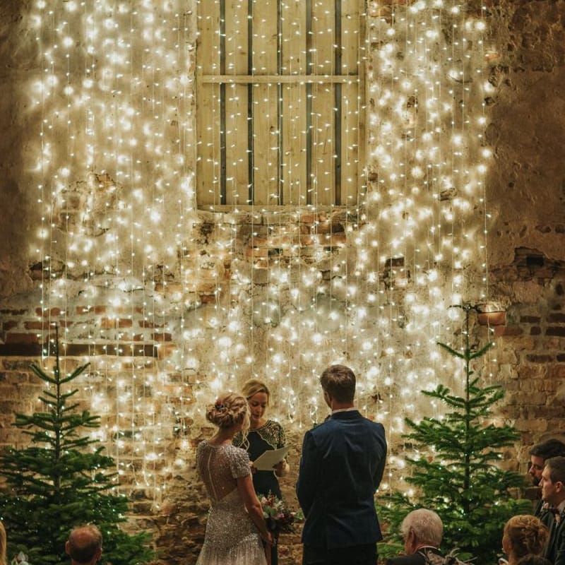 Bride and groom tie the knot at winter ceremony wearing a embellished wedding dress and navy velvet suit with fairy light curtain and Christmas trees