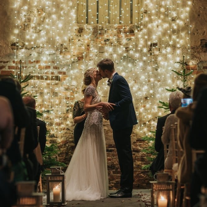 Bride and groom tie the knot at winter ceremony wearing a embellished wedding dress and navy velvet suit with fairy light curtain and Christmas trees