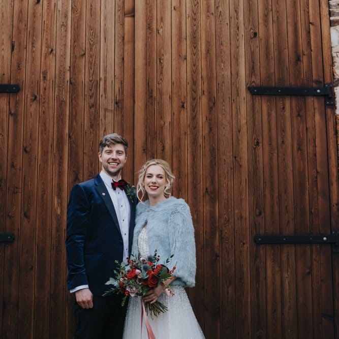 Bride wearing embellished wedding dress and jacket at winter wedding ceremony with red floral bouquet