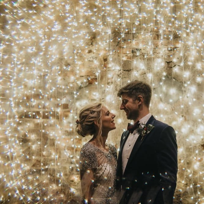 Bride and groom embrace with fairy light curtain at New Years eve wedding