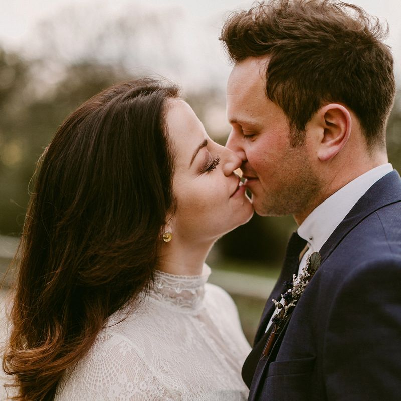 Bride in Homemade Wedding Dress with Lace Long Sleeves and High Neck and Groom in Navy Zara Suit Kissing