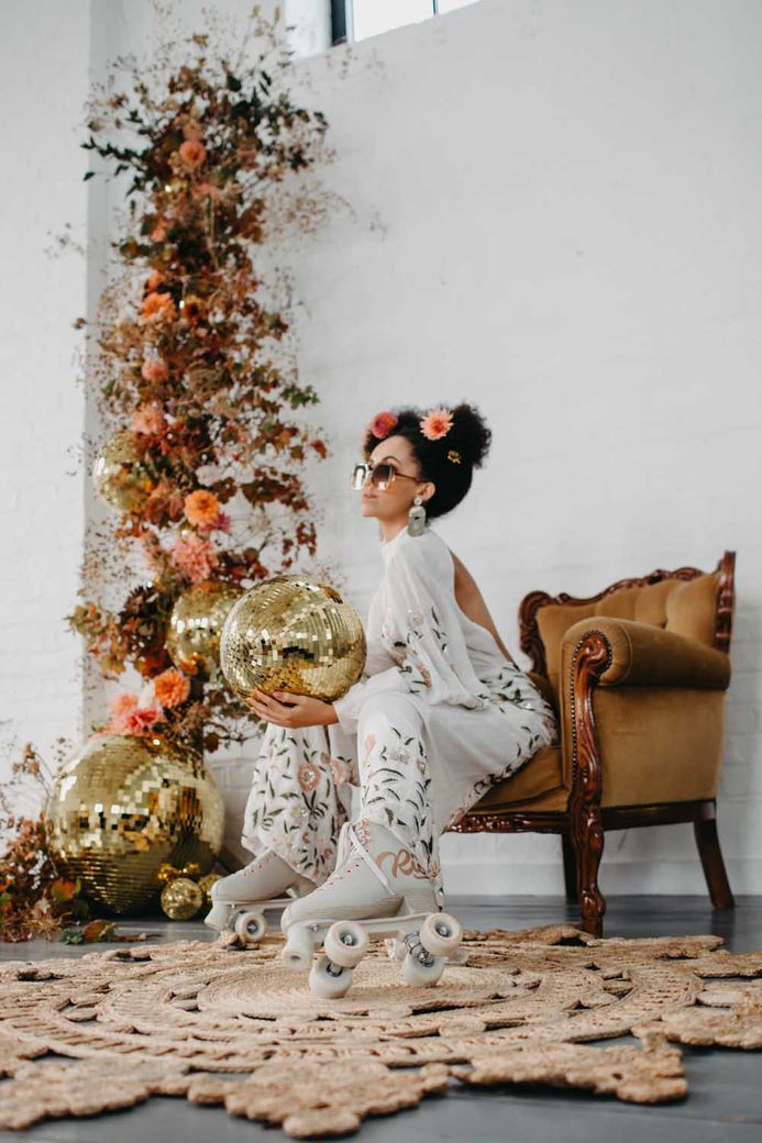 70s disco wedding inspiration with Black bride in a floral jumpsuit and roller skates sitting on an ornate couch holding a gold disco ball