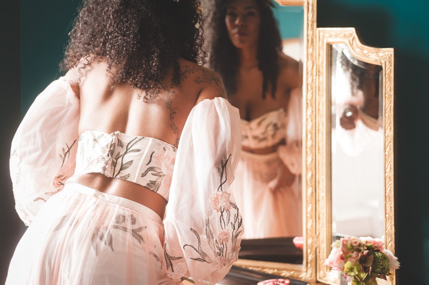 Black bride with long curly hair in blush pink bridal separates with appliqué detail looking in the mirror