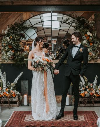 bride in a lace dress and groom in a black tuxedo at new Lake District wedding venue with oval micro back drop and colourful floral arrangements 