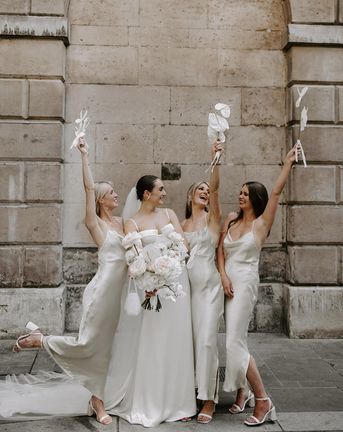 Bride holding white wedding bouquet with bridesmaids in white dresses.