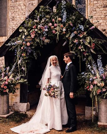 Bride in Jesus Peiro wedding dress and groom in suit stand in front of pink and blue wedding flowers display.