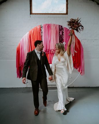 The bride and groom stand in front of pink, red and silver streamer decorations at the altar.