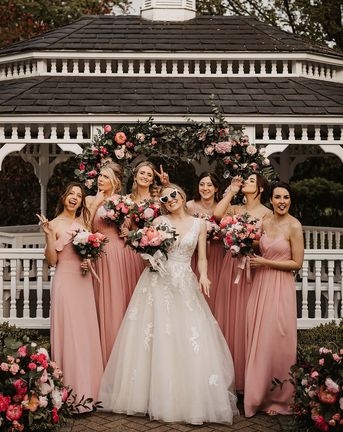 Bride with retro sunglasses with bridesmaids in pink dresses in front of a pink flower arch.