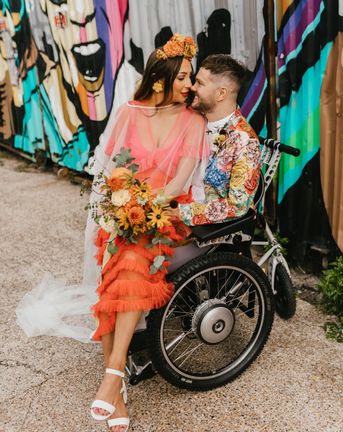 Bride in a coral wedding dress sitting on her grooms lap in a wheelchair for brightly coloured intimate elopement