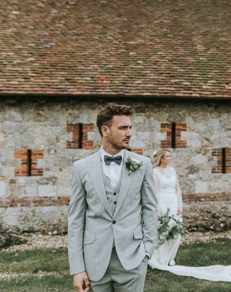 Portrait of the groom in a grey three-piece wedding suit with his bride in the background